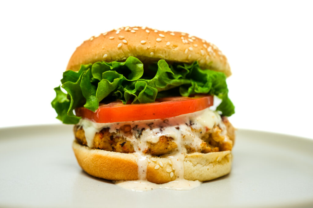 Grouper burger stacked with lettuce, tomato, and sauce on a sesame seed bun, served on a white plate.