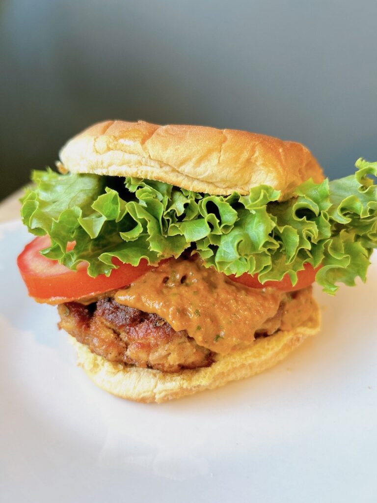 Alt text: "Juicy peri peri chicken burger on a toasted bun with lettuce, tomato and creamy sauce dripping down the sides, showcasing the vibrant orange-red Portuguese-African spicy sauce atop the golden-brown patty.