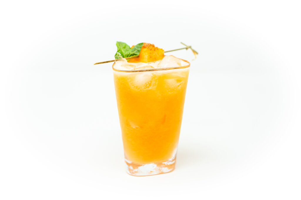 A golden-orange mezcal cocktail made with fresh cantaloupe and basil, served over ice and garnished with a basil sprig and cantaloupe cube.