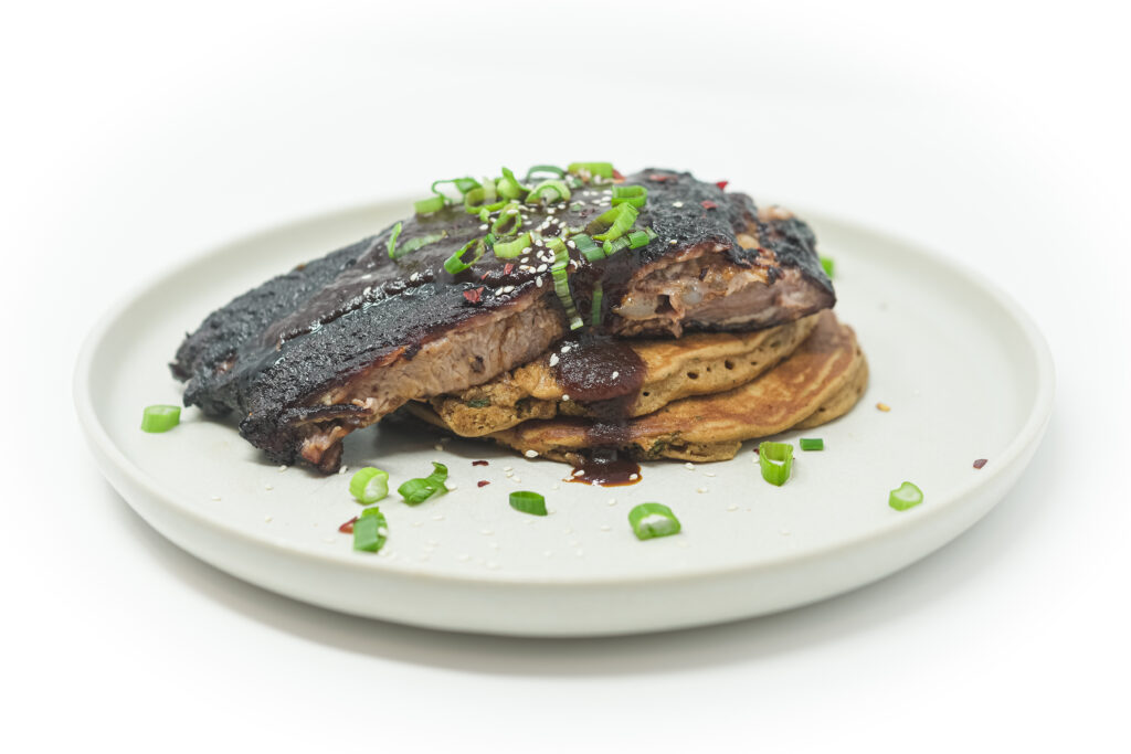 Slow-cooked pork spare ribs coated in a caramelized tomato-soy glaze, served on crispy cornmeal scallion pancakes, garnished with sliced scallions and sesame seeds.
