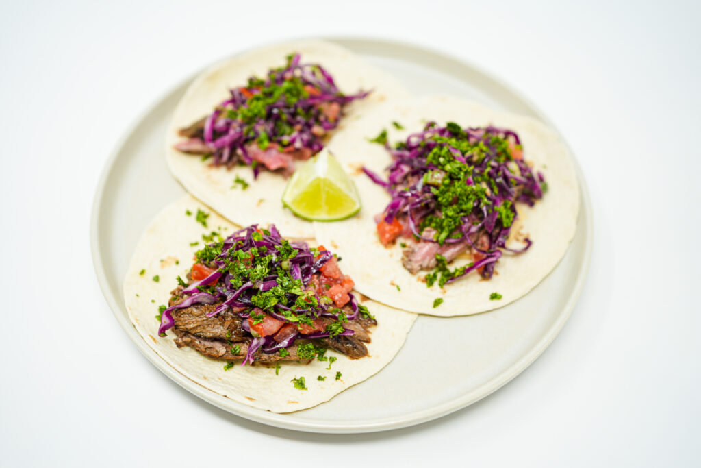 A vibrant image showing Cast-Iron Seared Flank Steak Tacos filled with juicy, caramelized flank steak, a colorful tangy red cabbage slaw and a creamy cheese sauce, all nestled in warm flour tortillas.