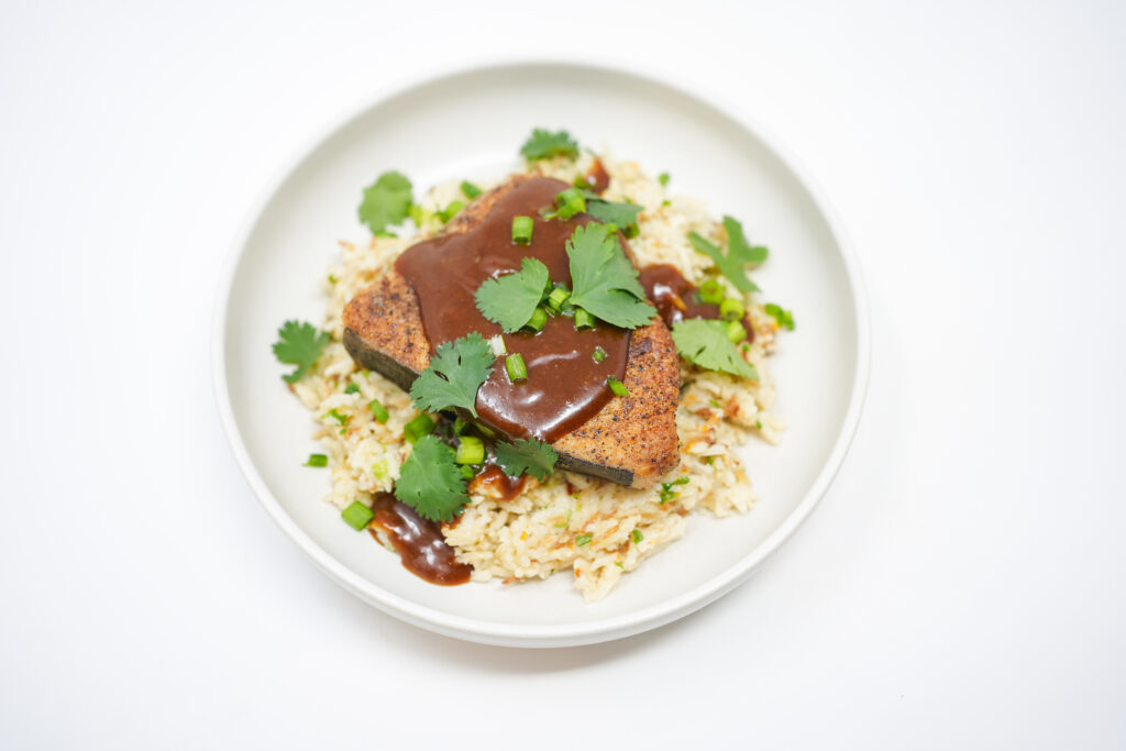A plate of pan-seared swordfish topped with a vibrant Thai-inspired mango sauce, served atop coconut caramelized rice, with fresh lime wedges on the side, showcasing the rich golden-brown fish and the colorful, tropical sauce.