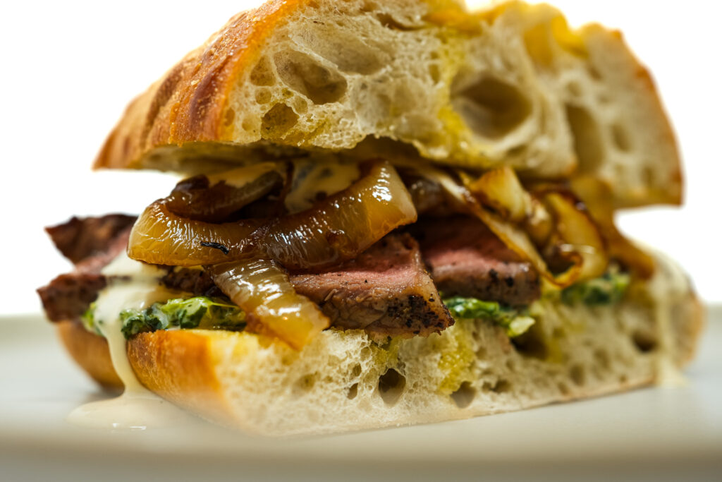 A ny strip steak sandwich with tender slices of seared steak on a toasted bun, topped with creamy spinach and caramelized onions, drizzled with steak sauce.