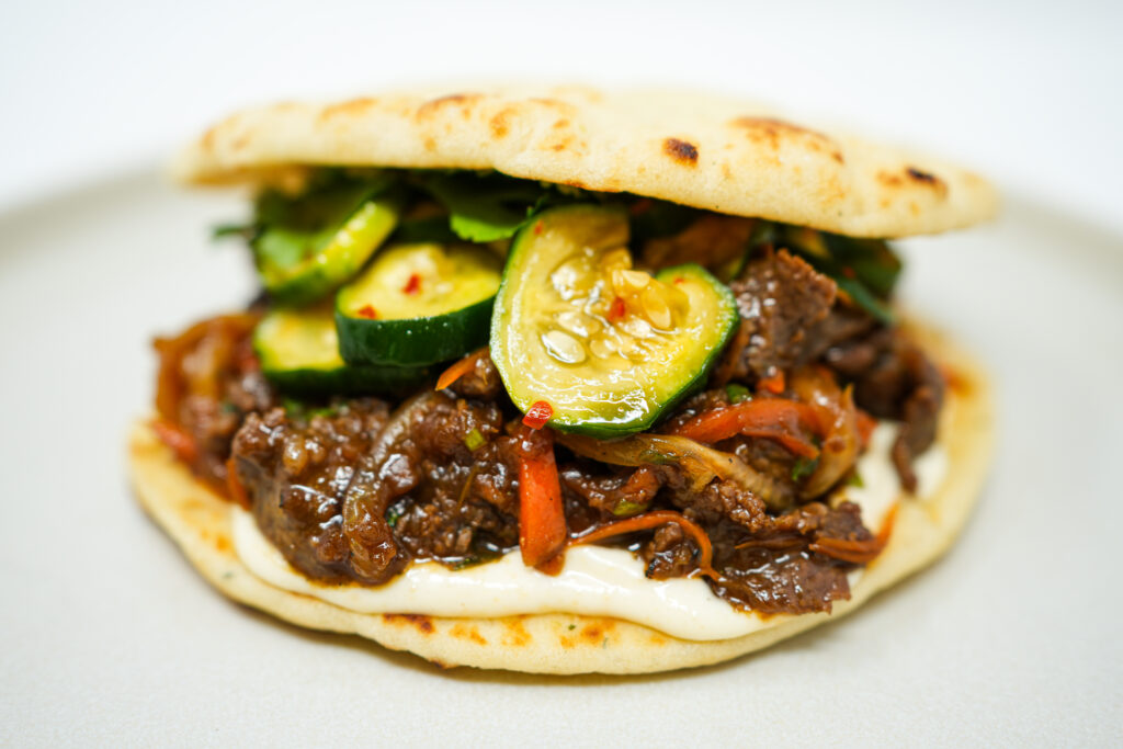 A Beef Bulgogi Sandwich on toasted naan bread, topped with spicy marinated cucumbers, a drizzle of garlic mayo, and fresh cilantro leaves, showcasing a delicious fusion of flavors.