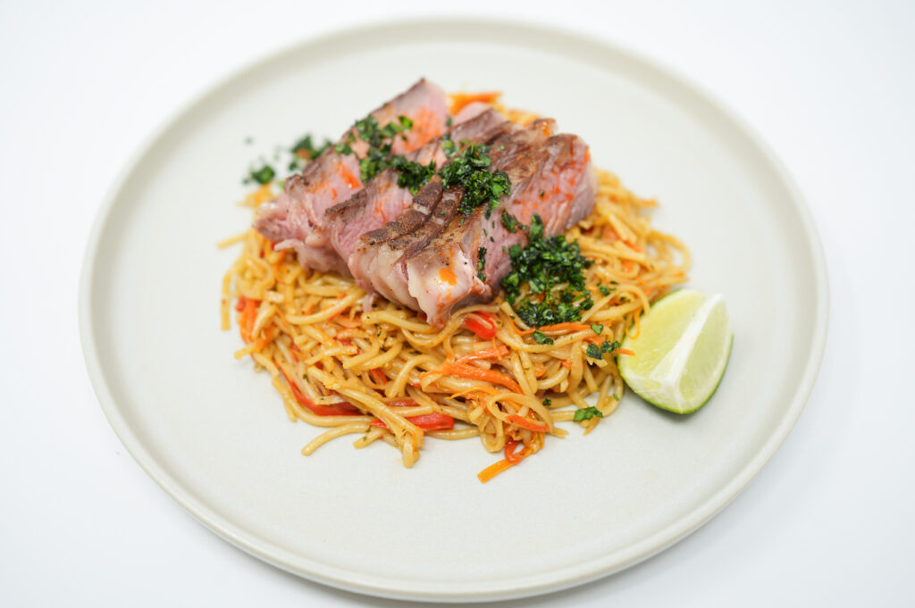 A tantalizing dish featuring reverse seared ribeye steak, thinly sliced and arranged atop a colorful stir-fry of udon noodles, red bell peppers, onions, and carrots, garnished with fresh cilantro and a drizzle of chili oil, blending Eastern and Western culinary styles in a visually appealing and flavorful presentation.