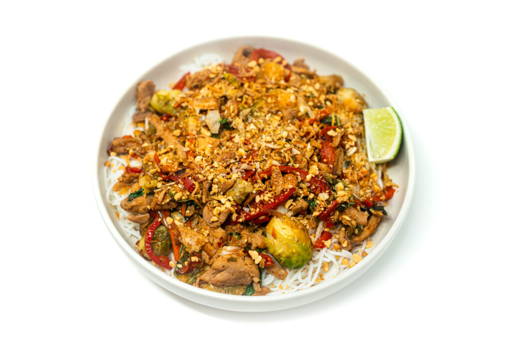 A vibrant plate of Thai Chili Chicken & Brussels Sprouts Stir-Fry, featuring golden seared chicken, crispy Brussels sprouts, red bell peppers, and Thai chilies, all coated in a glossy, umami-rich sauce and garnished with fresh basil, cilantro, and crushed peanuts.