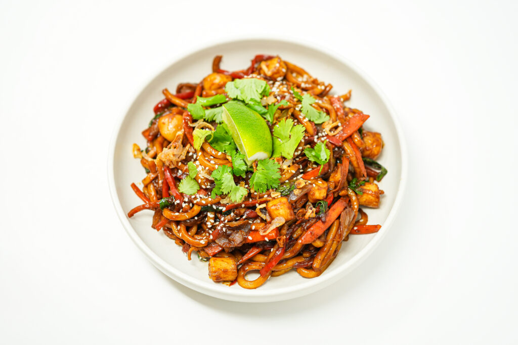 A serving of spicy scallop udon stir-fry with seared scallops, udon noodles, sautéed vegetables, fried shallots, and a garnish of sesame seeds and lime wedges.