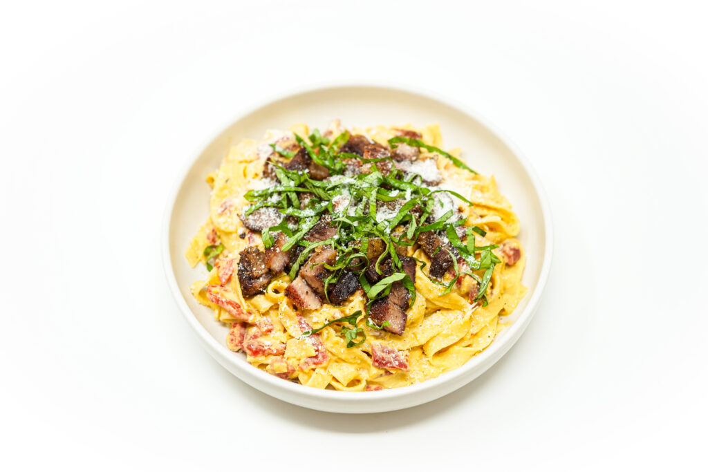 A plate of crispy pork belly pieces served over creamy corn-coated tagliatelle, with sautéed red bell peppers, fresh herbs, grated Parmesan, and lemon zest.