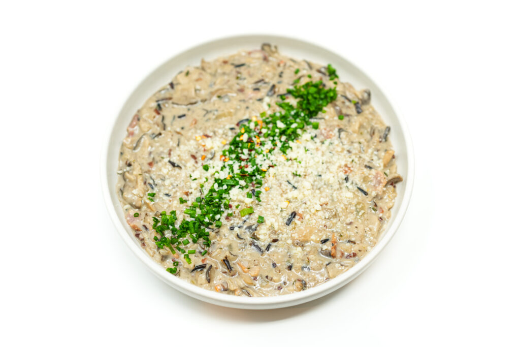 A bowl of Mezcal-Infused Creamy Mushroom and Wild Rice Soup, featuring golden-brown shiitake mushrooms, wild rice, and crispy bacon in a rich, creamy broth, garnished with chopped chives and Parmesan cheese.