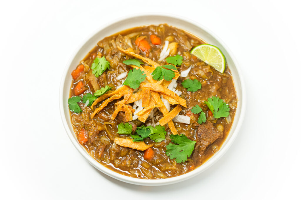 A bowl of Mexican Beef and Vegetable Stew (Caldo de Res) filled with tender beef chunks, carrots, potatoes, zucchini, corn, and cabbage in a flavorful broth, garnished with fresh cilantro, diced onions, and crispy tortilla strips.