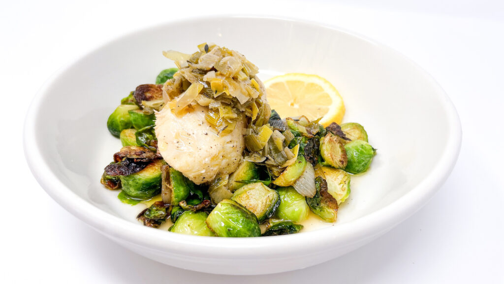 White wine poached cod served alongside rich leek confit and crispy sautéed Brussels sprouts, elegantly plated with a balance of soft, tender fish and crisp, golden vegetables.