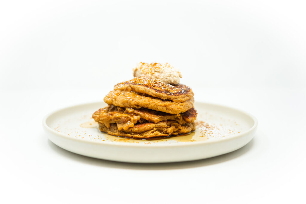A stack of fluffy Biscoff cookie pancakes is topped with creamy Biscoff cream cheese spread, drizzled with maple syrup, and garnished with crushed Biscoff cookies.