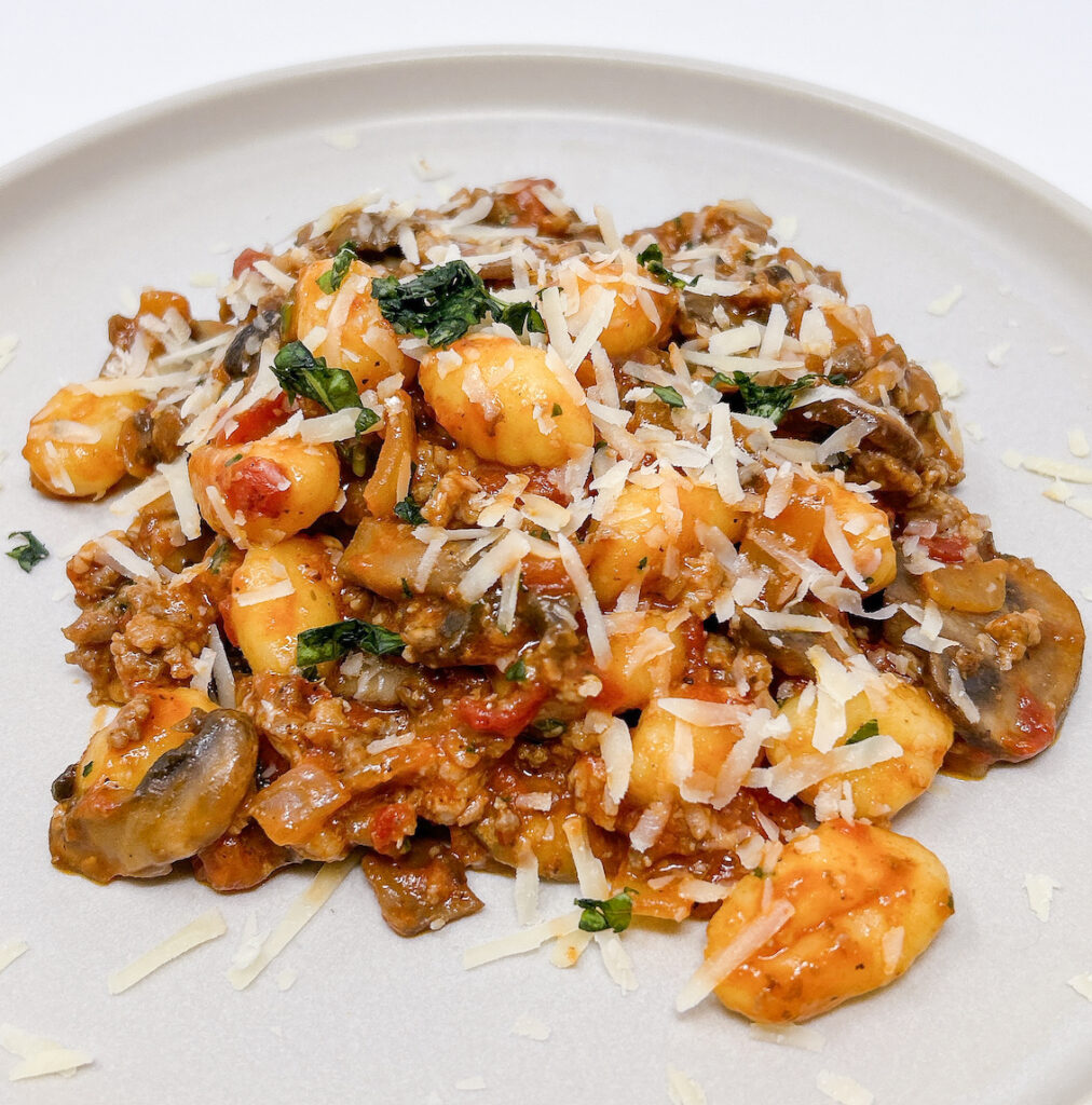 Plate of gnocchi with Italian sausage, sautéed mushrooms, and a rich tomato sauce, garnished with fresh basil and Parmesan cheese.