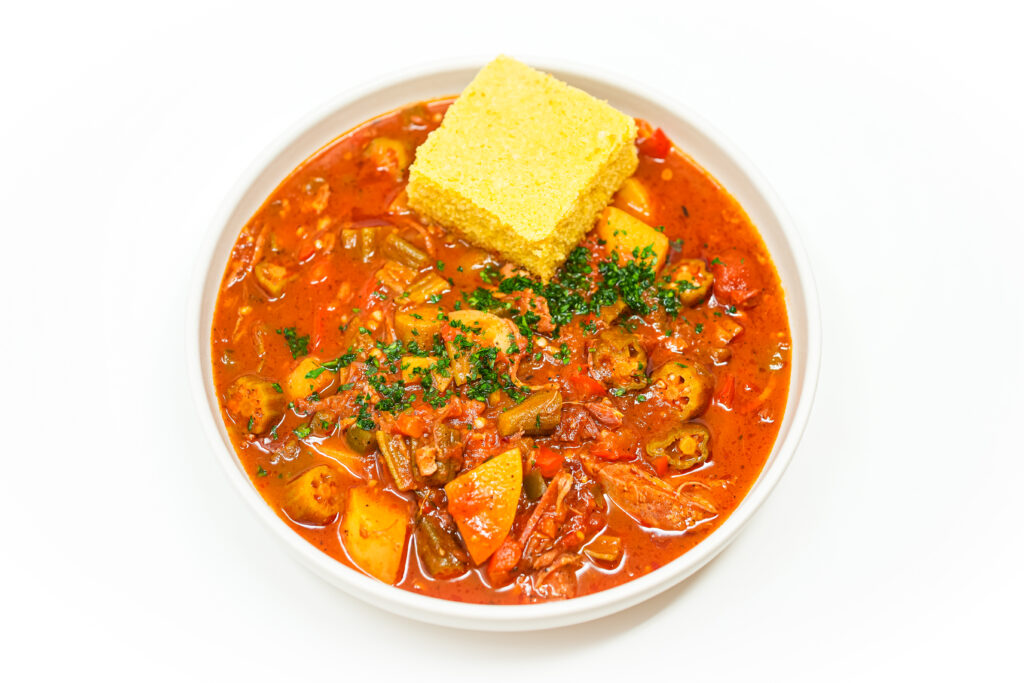 Bowl of ham and okra stew with tender vegetables and shredded ham, garnished with fresh parsley, alongside a slice of golden creamy cornbread.