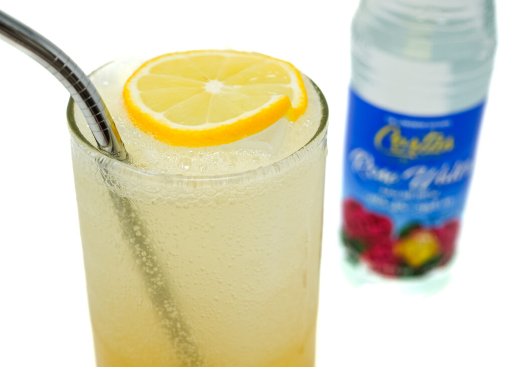 A tall glass of honey rose water vodka lemonade with a metal straw and lemon garnish, next to a bottle of Cortas Rose Water.