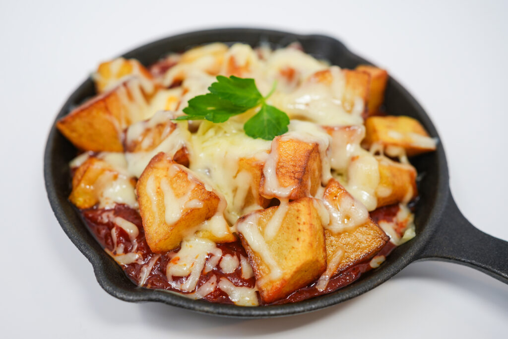 Roasted Red Pepper and Monterey Jack Cheese Patatas Bravas served in a cast iron skillet, featuring crispy potatoes topped with smoky roasted red pepper sauce and melted Monterey Jack cheese, creating a savory and flavorful dish with a rustic presentation