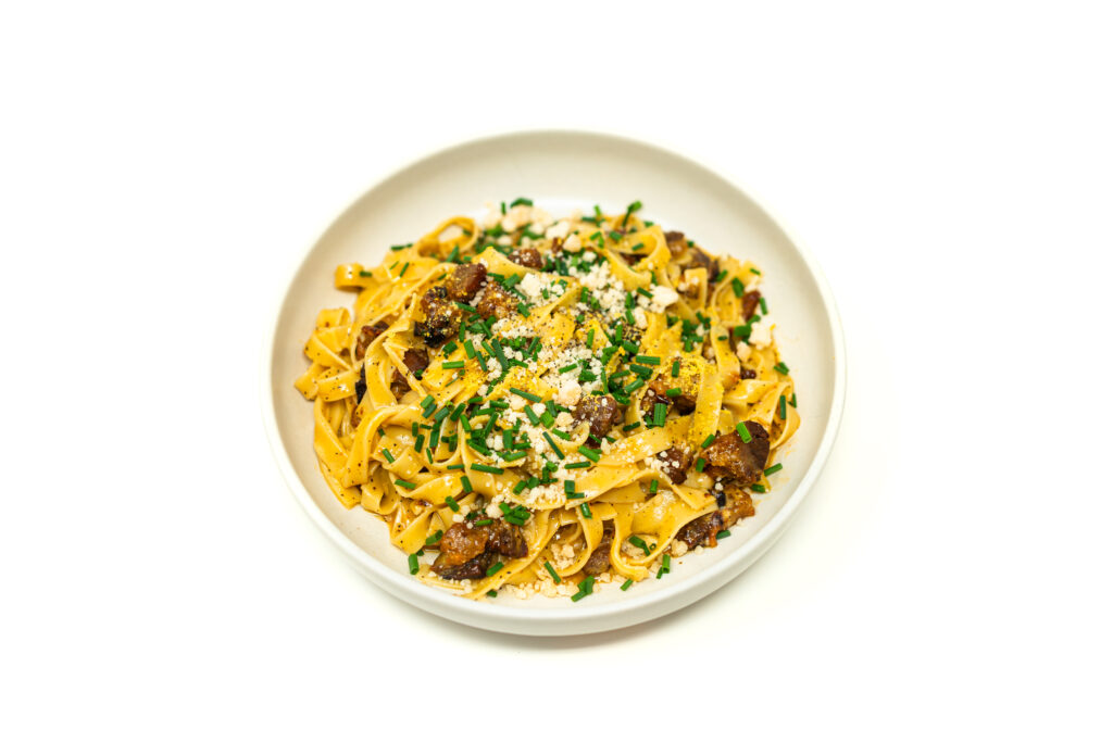 A bowl of miso pork belly carbonara garnished with fresh chives and grated parmesan cheese on a white background.