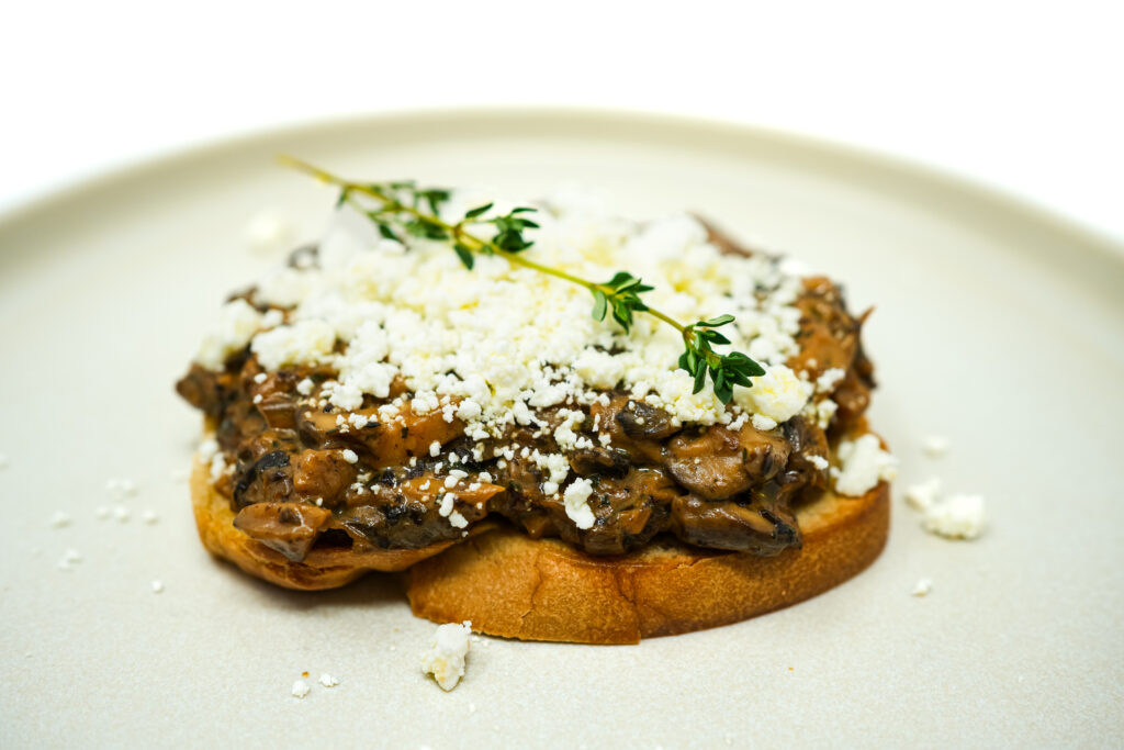 Shiitake and Oyster Mushroom Toast topped with sautéed mushrooms on crusty bread, garnished with fresh herbs, showcasing the earthy, rich flavors of the mushrooms against the golden-brown toast.