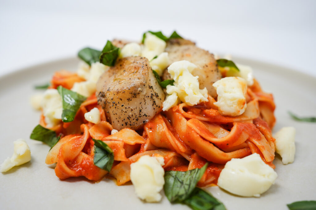 Tagliatelle al Pomodoro served with seared sea scallops and fresh mozzarella, featuring tender pasta in a rich tomato sauce, complemented by perfectly browned scallops and creamy mozzarella for a flavorful, elegant dish