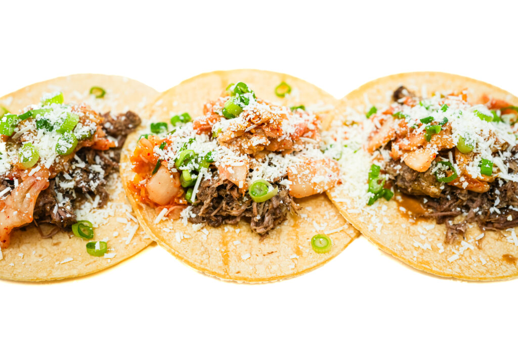 Three kimchi pork carnitas tacos on a plate, showcasing juicy pork topped with vibrant, spicy kimchi, nestled in soft tortillas, ready to be enjoyed.