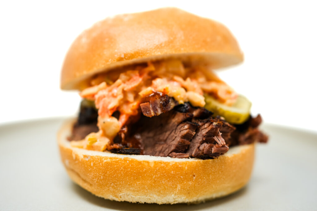 A delectable sweet and tangy brisket sandwich on ciabatta bread, layered with juicy beef and vibrant kimchi coleslaw, creating a colorful and appetizing meal.