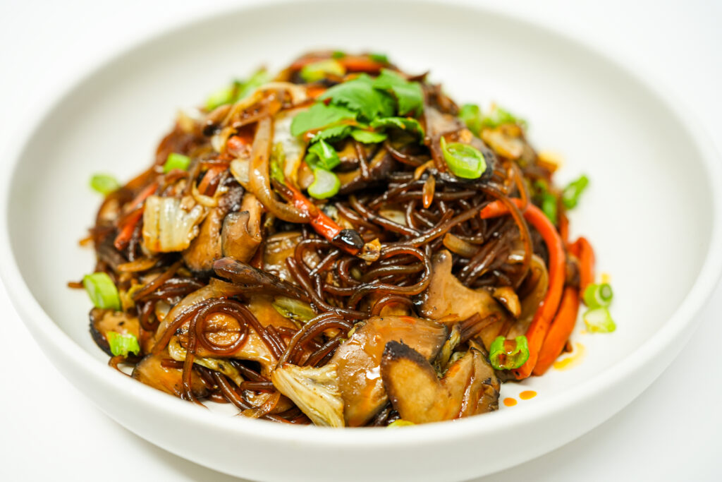 Plate of Shiitake Mushroom and Napa Cabbage Japchae, featuring glossy, stir-fried glass noodles mixed with tender shiitake mushrooms, crisp Napa cabbage, and vibrant vegetables, garnished with green onions.