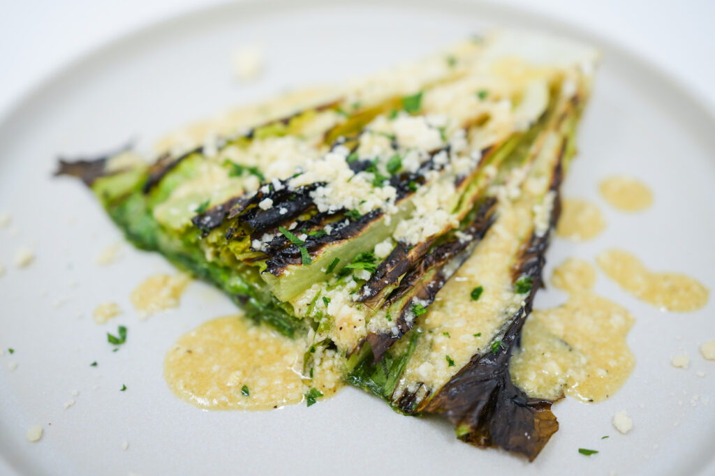 A smoky charred romaine lettuce salad drizzled with zesty lemon parmesan dressing, topped with delicate shavings of Parmesan cheese, presented on a modern plate.