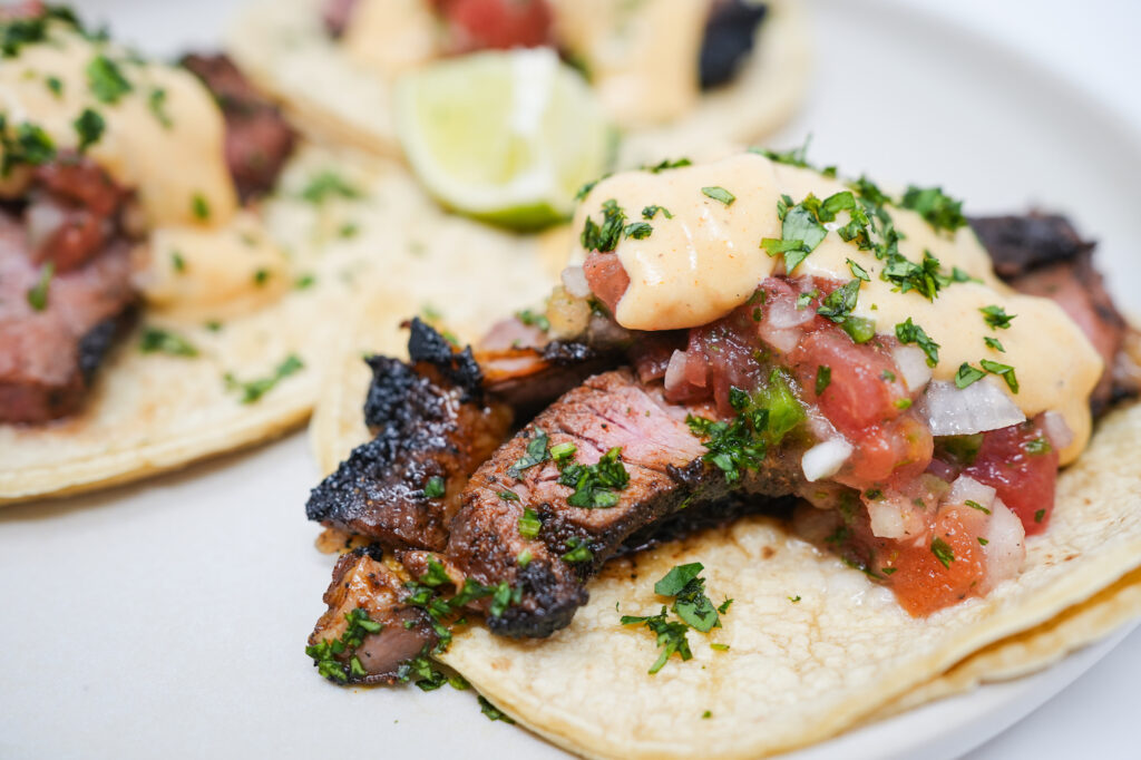 Coffee-Ancho Sirloin Steak Tacos drizzled with creamy Pepper Jack Sauce, nestled in soft tortillas and topped with vibrant, fresh tomato salsa and a sprinkle of cilantro, embodying a perfect blend of smoky, spicy, and tangy flavors.