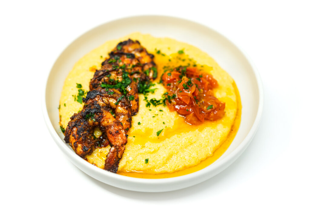A colorful and appetizing plate of Spicy Grilled Shrimp and Grits featuring succulent, charred shrimp atop creamy, golden grits, complemented by a vibrant cherry tomato confit garnished with fresh herbs.