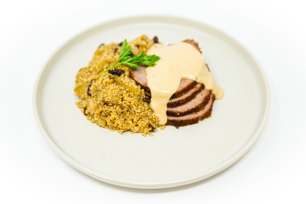 A succulent, spice-crusted eye round roast beef, sliced and generously drizzled with creamy lemon-tahini sauce, accompanied by a fluffy side of couscous, all presented on a white platter showcasing Moroccan-inspired culinary elegance.
