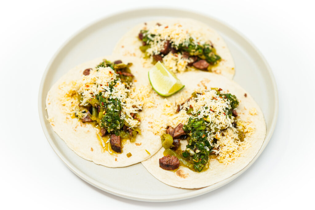 A vibrant image of Grilled Chorizo and Cubanelle Pepper Tacos, featuring juicy, charred chorizo and sliced green Cubanelle peppers topped with melted Monterey Jack cheese and a drizzle of bright green chimichurri sauce, all nestled in a lightly toasted flour tortilla.