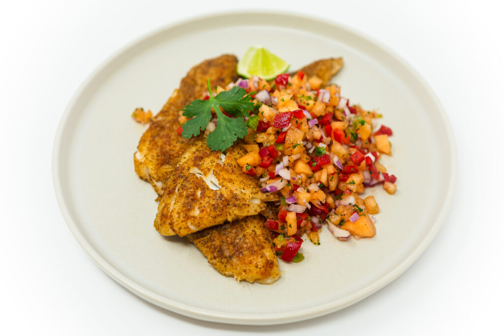 A plate featuring pan-fried haddock topped with a colorful cantaloupe salsa, highlighting the crispy golden exterior of the fish contrasted against the bright orange and green hues of the diced cantaloupe, red bell pepper, and fresh cilantro.