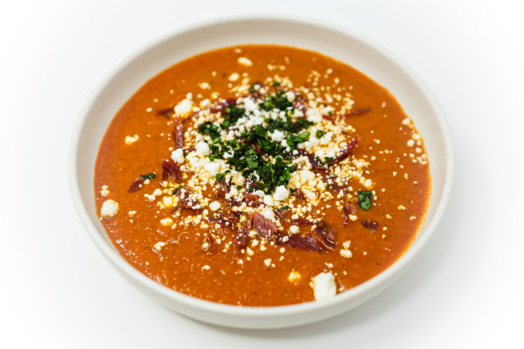 A bowl of Roasted Tomato, Cantaloupe, and Pancetta Soup, featuring a creamy blend of vibrant orange soup topped with golden crispy pancetta, crumbled goat cheese, and a sprinkle of fresh green parsley.
