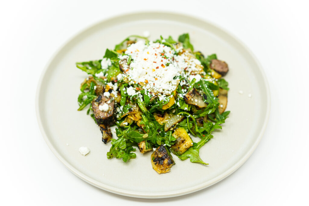 A vibrant image of Grilled Sausage and Summer Squash Salad featuring slices of smoky grilled Italian sausage, charred zucchini and yellow squash, and crisp arugula leaves, all tossed together and sprinkled with crumbled goat cheese. The salad is drizzled with a lemon herb vinaigrette and garnished with red pepper flakes, adding a subtle hint of spice. The colors and textures create a visually appealing and appetizing dish.