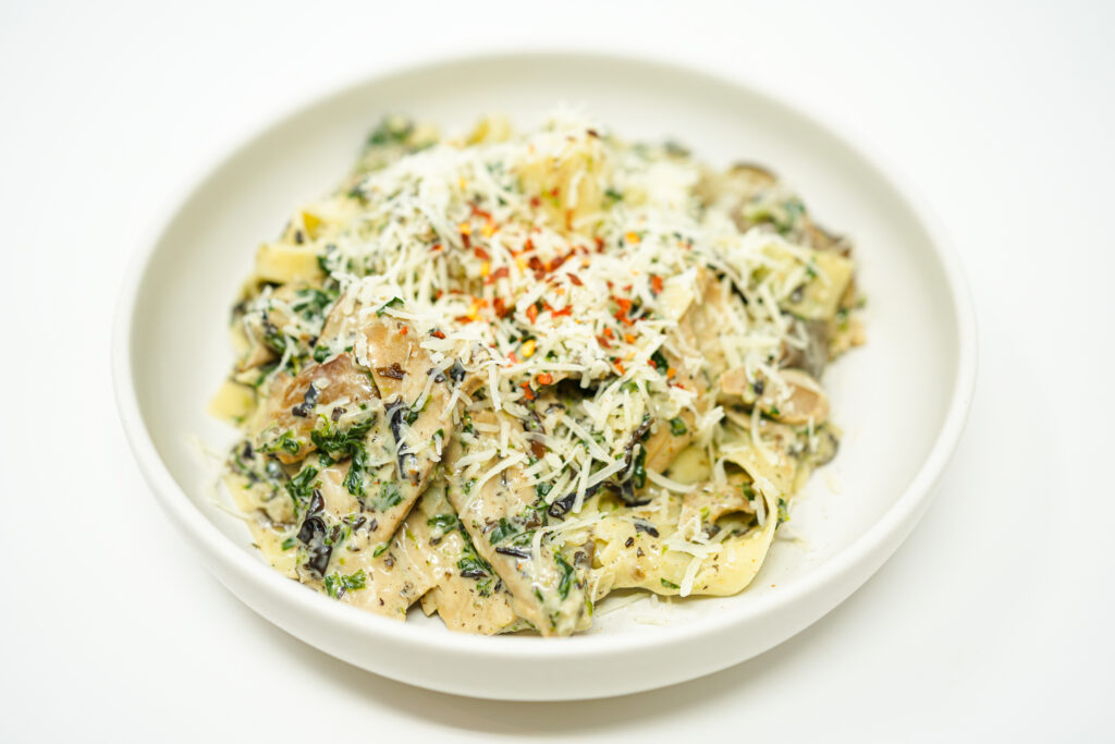 A plate of Creamy Spinach and Wild Mushroom Pappardelle, showcasing wide ribbons of pasta coated in a rich, creamy sauce with vibrant green spinach and golden-brown mushrooms, sprinkled with red pepper flakes for a touch of warmth.