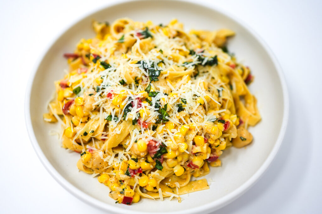 A plate of Creamy Corn and Bay Scallop Fettuccine, with plump scallops and golden corn kernels nestled in ribbons of pasta, draped in a silky, lemony Parmesan sauce and garnished with fresh basil.