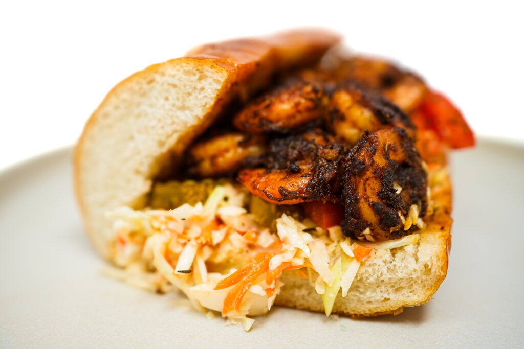 A Blackened Shrimp and Creamy Coleslaw Po'Boy sandwich with spicy shrimp, creamy coleslaw, bread and butter pickles, and fresh tomato slices in a ciabatta roll.