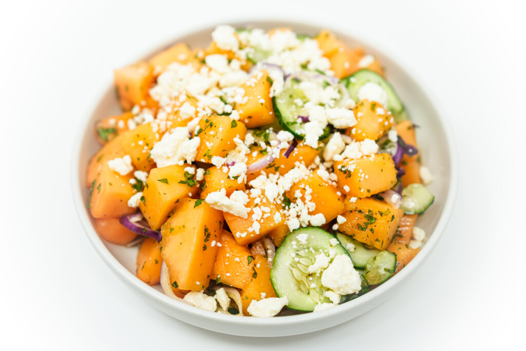 Fresh Mediterranean cantaloupe salad served in a white bowl, featuring bright orange melon cubes, sliced cucumber, crumbled feta cheese, and fresh herbs. The colorful summer dish showcases contrasting textures and vibrant ingredients.