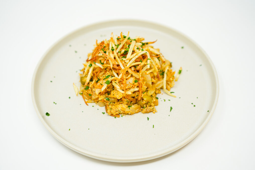 A vibrant and appetizing dish of Bacalhau à Brás with Olives and Paprika, featuring flaked salt cod mixed with golden, crispy julienned potatoes, scattered green and black olives, and a sprinkle of bright red paprika. The dish is garnished with fresh green parsley, adding a burst of color and freshness, served on a white ceramic plate.