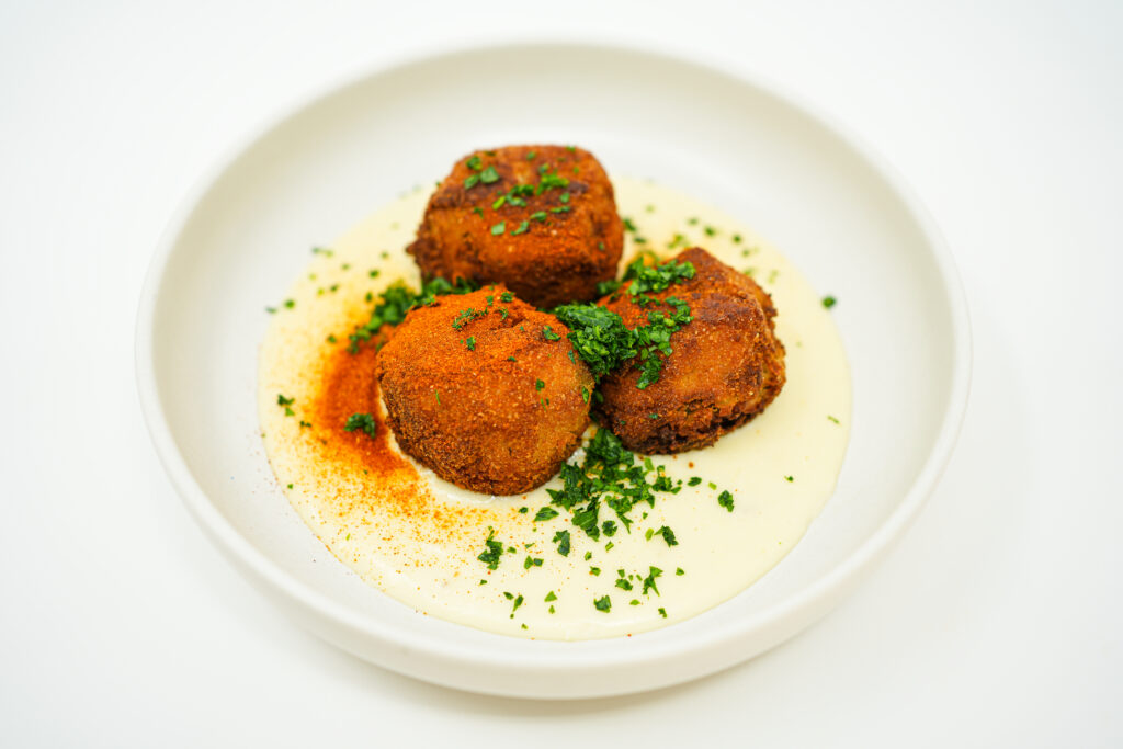 A plate of Chicken and Potato Croquettes with Gruyere Cheese Sauce, featuring crispy, golden croquettes resting on a bed of creamy cheese sauce, garnished with chopped parsley and paprika.