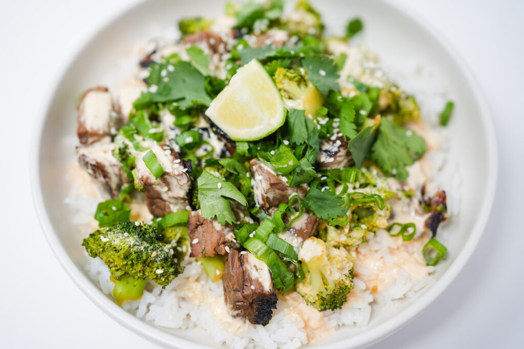 Asian-Inspired Skirt Steak and Broccoli Bowls with Mango Aioli: A vibrant bowl featuring sliced marinated skirt steak, tender steamed broccoli, and fluffy jasmine rice, drizzled with a savory-sweet sauce and creamy mango aioli, garnished with fresh green onions, cilantro, and sesame seeds.
