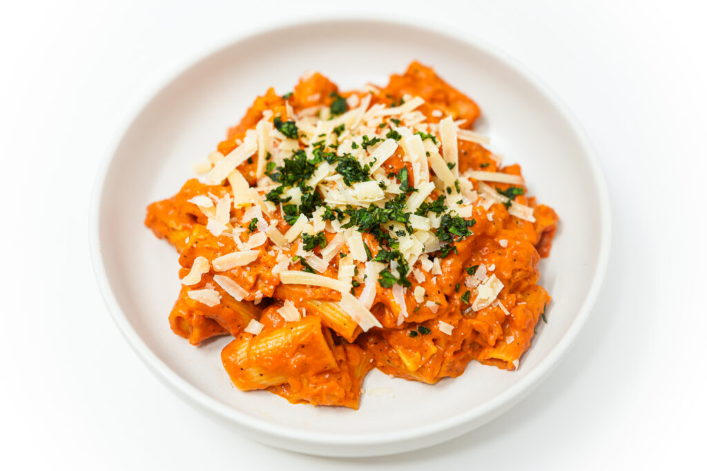 A plate of Garlic Butter Vodka Rigatoni, garnished with freshly grated Parmesan cheese and vibrant green basil leaves. The rigatoni pasta is coated in a rich, creamy sauce, with visible specks of garlic and a hint of red from the pureed tomatoes.