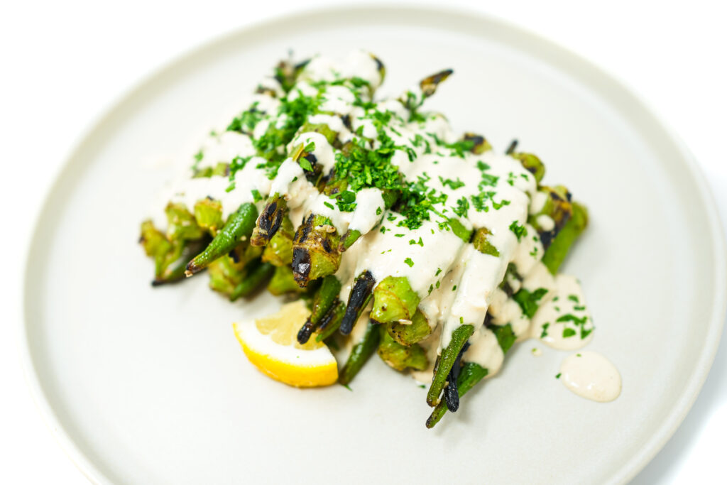 A plate of Garlic Lemon Grilled Okra drizzled with creamy tahini dressing, garnished with fresh parsley and lemon zest. The okra is slightly charred and arranged on a white plate, showcasing its vibrant green color and the smooth, rich tahini sauce.