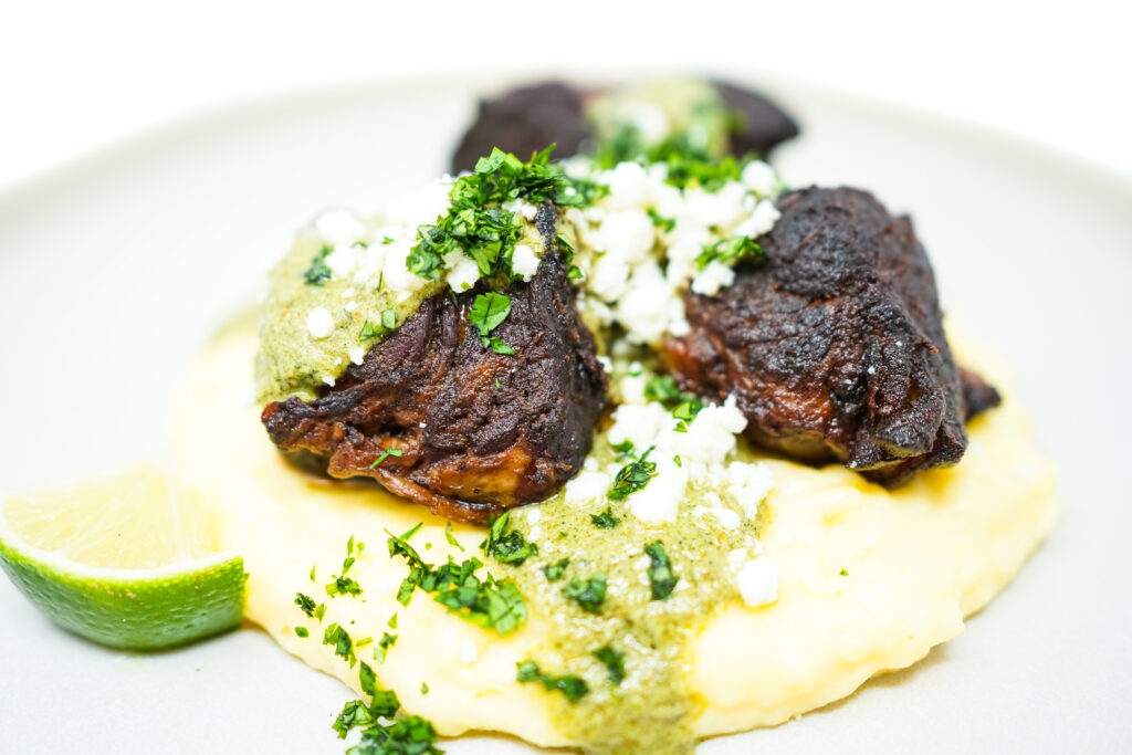 A plate of Pork Carnitas with Green Mole and Mashed Potatoes, featuring tender, crispy pork pieces on a bed of creamy mashed potatoes, drizzled with vibrant green mole sauce, and topped with crumbled queso fresco and fresh cilantro, garnished with lime wedges.
