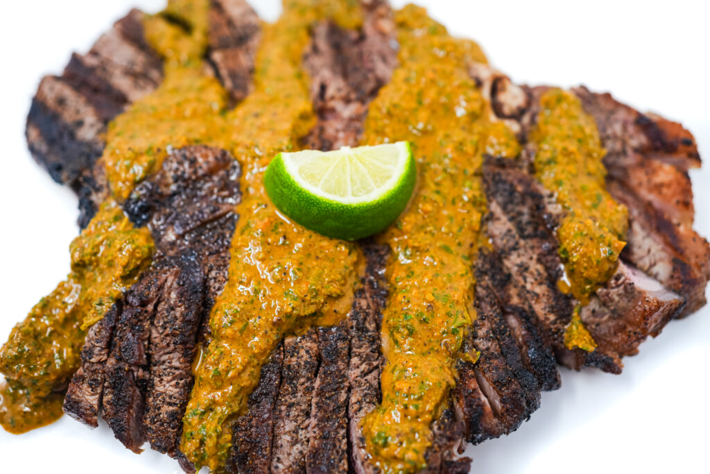 A perfectly grilled porterhouse steak topped with a vibrant, jerk-inspired chimichurri sauce made from fresh herbs, garlic, hot peppers, and warm spices, served on a white plate.