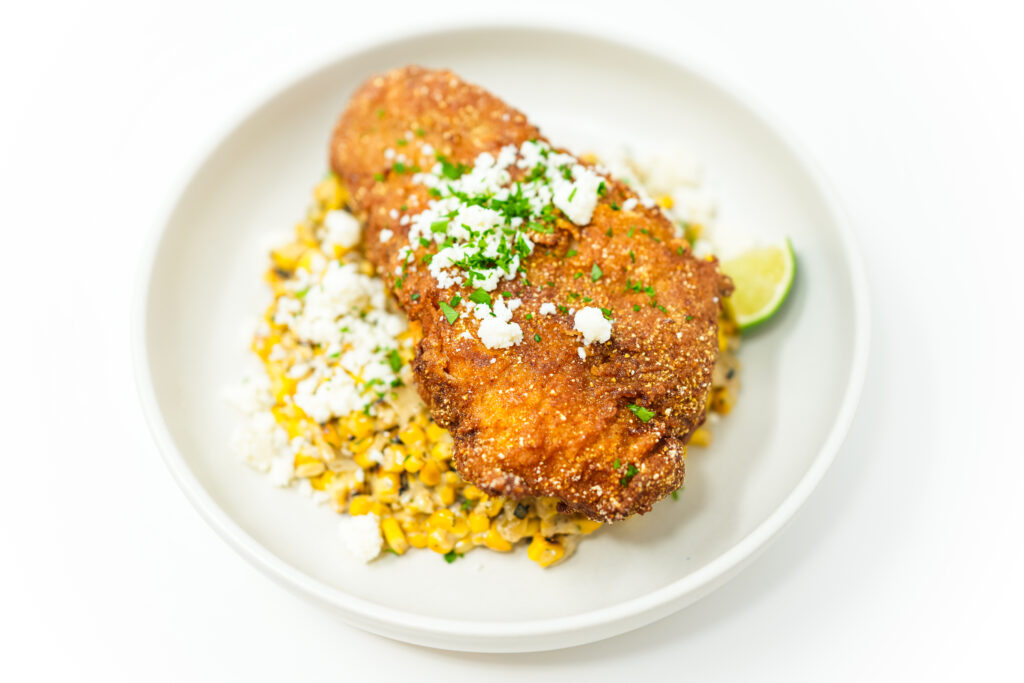A plate of Southern Fried Catfish with Spicy Esquites, featuring golden, crispy catfish fillets alongside creamy, charred corn topped with crumbled cotija cheese, fresh cilantro, and lime wedges.
