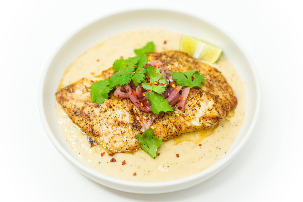 A beautifully plated dish of Pan-Seared Corvina with Coconut Lime Sauce, featuring golden-brown fish fillets atop a creamy, tangy sauce, garnished with vibrant pickled onions, fresh cilantro, lime wedges, and a sprinkle of red pepper flakes.