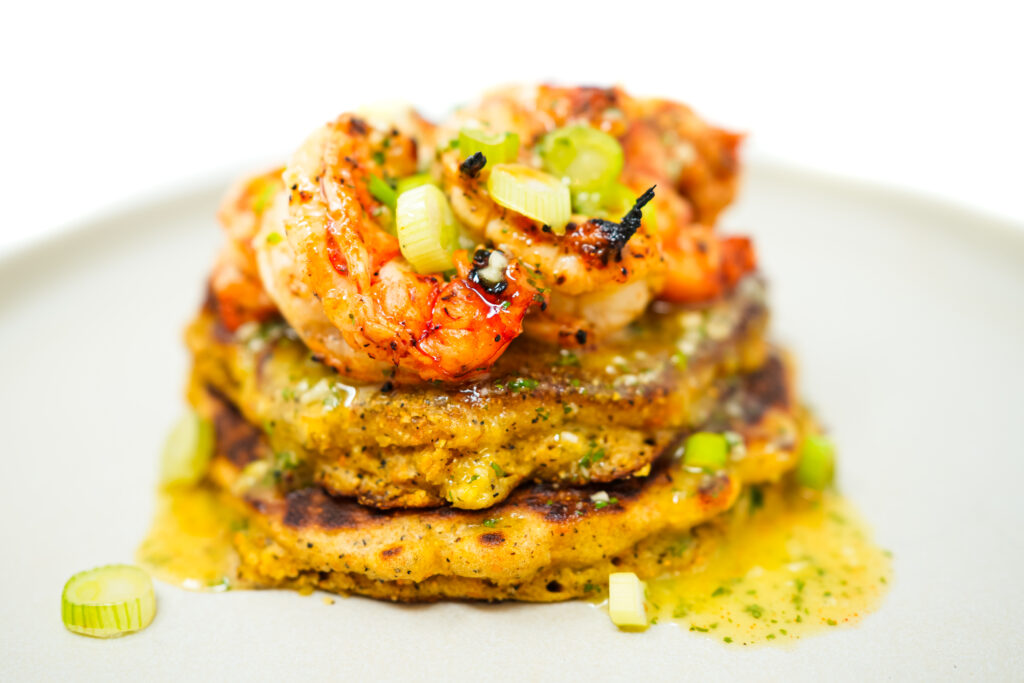 A plate of Citrus Grilled Shrimp skewers, served with golden cornmeal pancakes, a grilled lemon half, and drizzled with creamy cilantro-lime aioli.