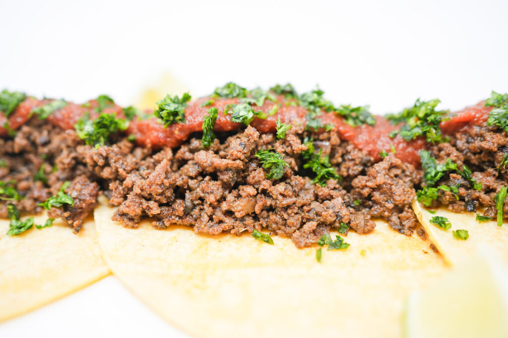 A plate of Ground Beef and Mushroom Duxelles Tacos filled with savory beef and mushroom mixture, topped with vibrant roasted tomato and pasilla pepper salsa, garnished with fresh cilantro, and served with lime wedges on warm flour tortillas.