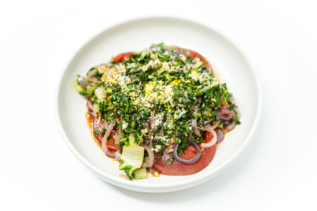A vibrant plate of Purple Tomato and Sautéed Green Chard Salad, featuring thinly sliced purple tomatoes topped with sautéed green chard and purple onions, drizzled with a tangy vinaigrette and garnished with lemon zest, grated Parmesan cheese, and fresh basil.